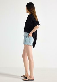 Svart långärmad tröja instoppad i ljusblå denimshorts med fransig nederkant. Modellen bär svarta flip-flops och står mot en neutral bakgrund.