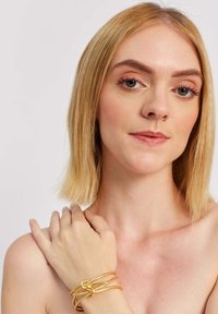 Złota bransoletka o splecionym wzornictwie, z gładkiego, błyszczącego metalu i elastycznym kształtem, noszona na nagim nadgarstku.