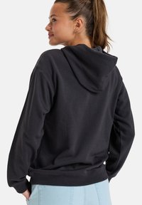 Schwarzer Kapuzenpullover aus strukturiertem Material, mit lockerer Passform und geripptem Saum. Das Modell trägt eine hellblaue Jeans.