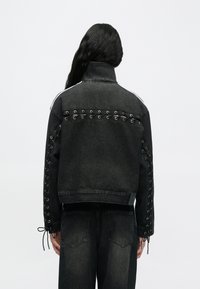 Μαύρο τζιν μπουφάν με δέσιμο με κορδόνια στα μανίκια και στην πλάτη. Διαθέτει ψηλό γιακά και αντίθετη ραφή κατά μήκος των ώμων.