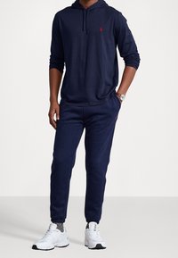 Námořnická modrá mikina s kapucí a odpovídající souprava tepláků vyrobená z měkké látky; obsahuje červené logo na hrudi a elastické manžety na kotnících.