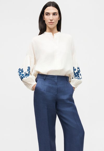Γυναίκα με κρεμ μπλούζα με μπλε ανθιστή κέντημα στα μανίκια και μπλε παντελόνι με φαρδιά μπατζάκια και ψηλή μέση, τα χέρια στις τσέπες, ουδέτερο φόντο.