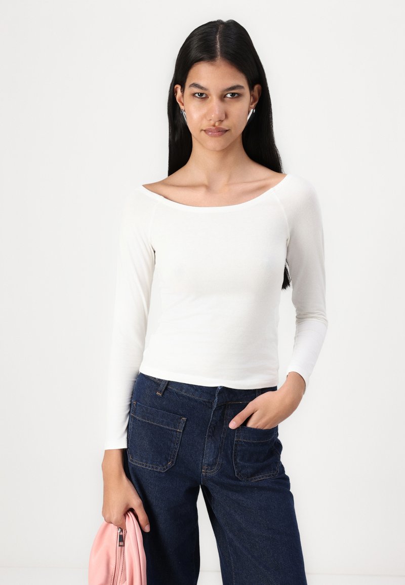 Top bianco a maniche lunghe con scollo ampio, abbinato a jeans blu scuro. Il modello tiene una borsa rosa. Design minimalista e texture liscia.