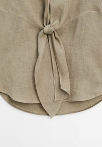 Tissu en lin beige noué en un noeud simple avec des extrémités libres dépassant d'un ourlet arrondi sur un fond blanc.