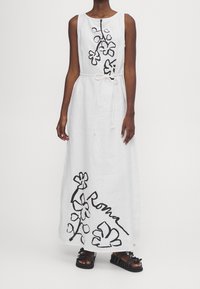 Valkoinen pellavainen maxi-mekko, jossa on musta kukkakuosi ja "Roma"-teksti. Mekossa on pyöreä pääntie, hihaton leikkaus ja kiristettävä vyötärö.