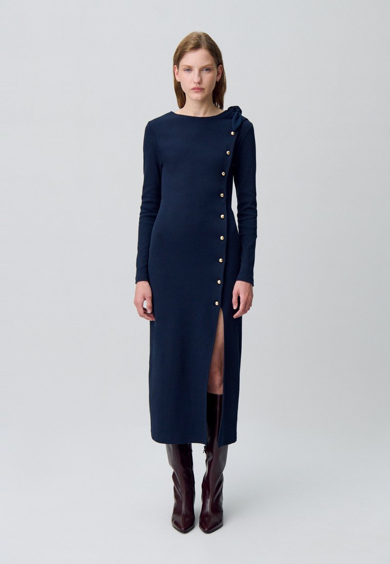 Robe à manches longues bleu marine avec une fente latérale, ornée de boutons dorés et d'un nœud sur l'épaule gauche. Texture de tissu lisse.