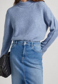 Pull en laine bleu clair avec un col montant côtelé, associé à une jupe en denim bleu moyen. Le modèle tient un sac à main noir matelassé.