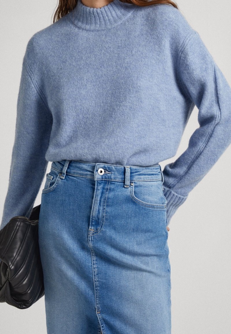 Pull en laine bleu clair avec un col montant côtelé, associé à une jupe en denim bleu moyen. Le modèle tient un sac à main noir matelassé.