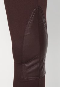 Brązowe legginsy z gładką skórzaną łatą na kolanie, o dopasowanym kroju i fakturowej powierzchni dla dodatkowego wykończenia.