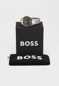 Ατσάλινο ρολόι με μαύρο καντράν, ασημένιο βραχιόλι και κόκκινες, κίτρινες και μπλε λεπτομέρειες; παρατίθεται σε μια μαύρη θήκη και υφασμάτινη τσάντα.