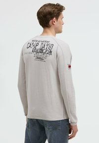 Hellgraues langärmliges Shirt mit bedruckten Grafiken auf dem Rücken, Raglanärmeln und Rundhalsausschnitt. Der Stoff wirkt weich mit leichter Struktur.