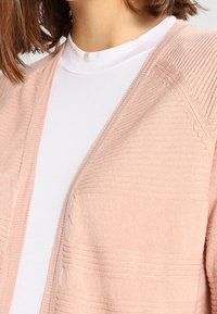 Cardigan rosa chiaro con texture a coste, dotato di due tasche frontali e una vestibilità rilassata, indossato sopra un dolcevita bianco.