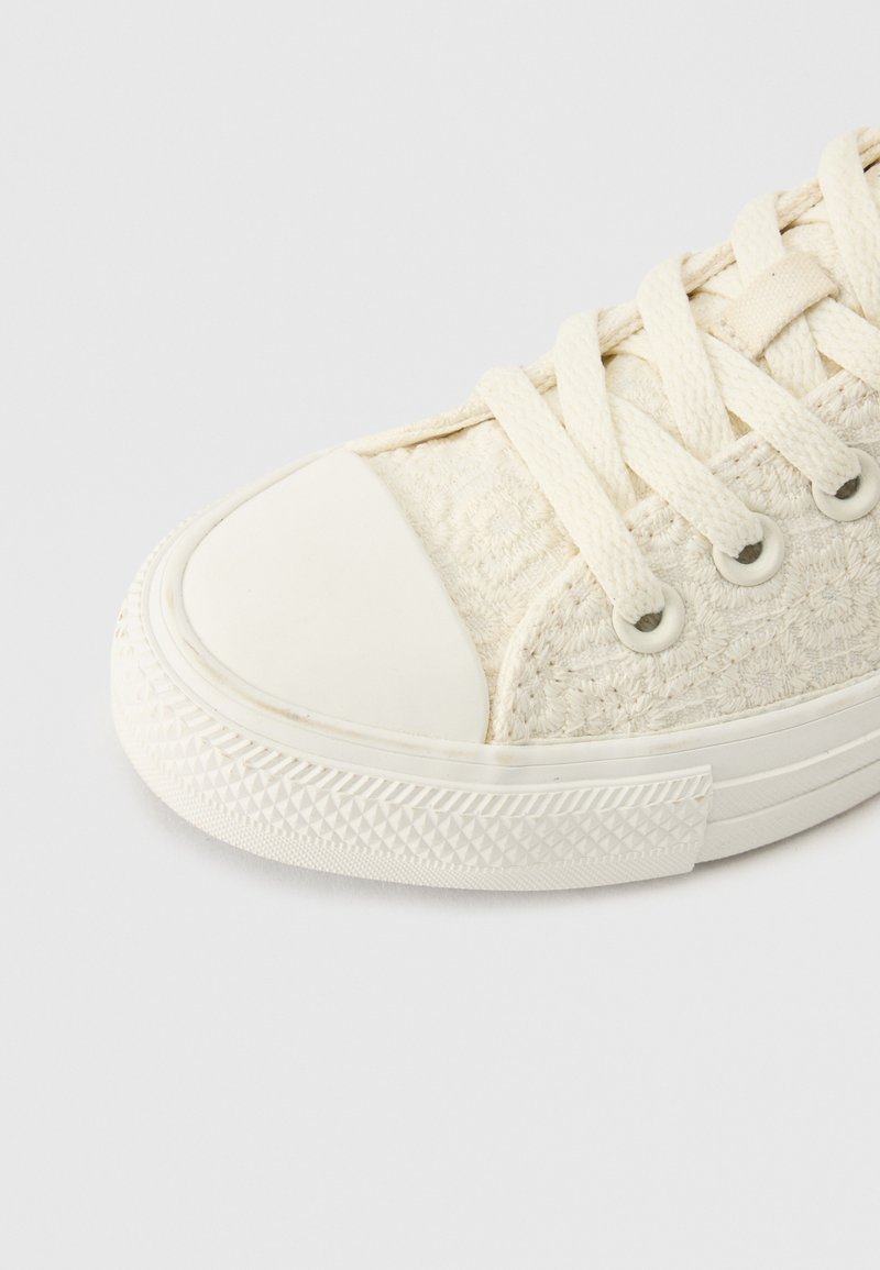 Primo piano di una sneaker in tela color crema con punta in gomma, suola texturizzata e lacci spessi intrecciati attraverso occhielli metallici.