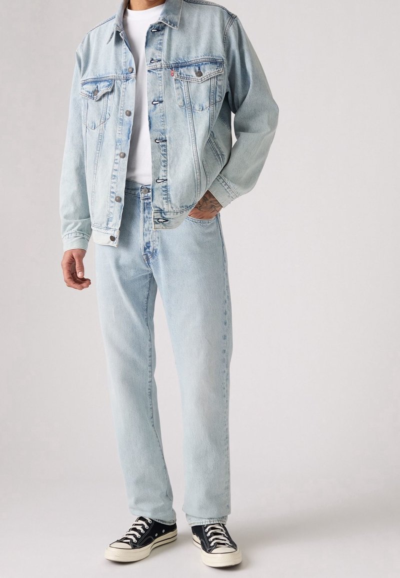 Homme portant une veste en jean bleu clair et un jean avec un t-shirt blanc et des baskets basses noires, debout devant un fond uni.