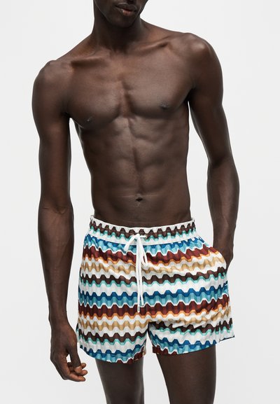 Homme torse nu à la peau foncée portant un short coloré à motif zigzag avec un cordon blanc, posant avec une main dans la poche sur un fond uni.