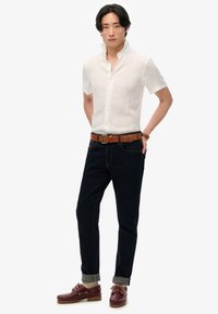 Biała lniana koszula z guzikami, ciemne dżinsy z żółtymi szwami, brązowy skórzany pasek oraz bordowe buty żeglarskie z jasnobrązowymi sznurówkami i gumową podeszwą.