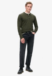 Donkergroen langarm henley-shirt met drie knopen, gecombineerd met donkerdenim jeans, opgerolde mouwen en zwarte veterschoenen.