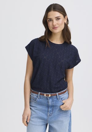 Młoda kobieta z brązowymi włosami, ubrana w granatową koronkową bluzkę, jasnoniebieskie dżinsy i brązowy pasek, stojąca z jedną ręką w kieszeni na białym tle.