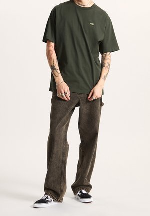 Personne portant un t-shirt Vans vert foncé, un jean marron ample, des baskets Vans noires, des tatouages visibles sur les bras, ainsi que des bagues et bracelets argentés.