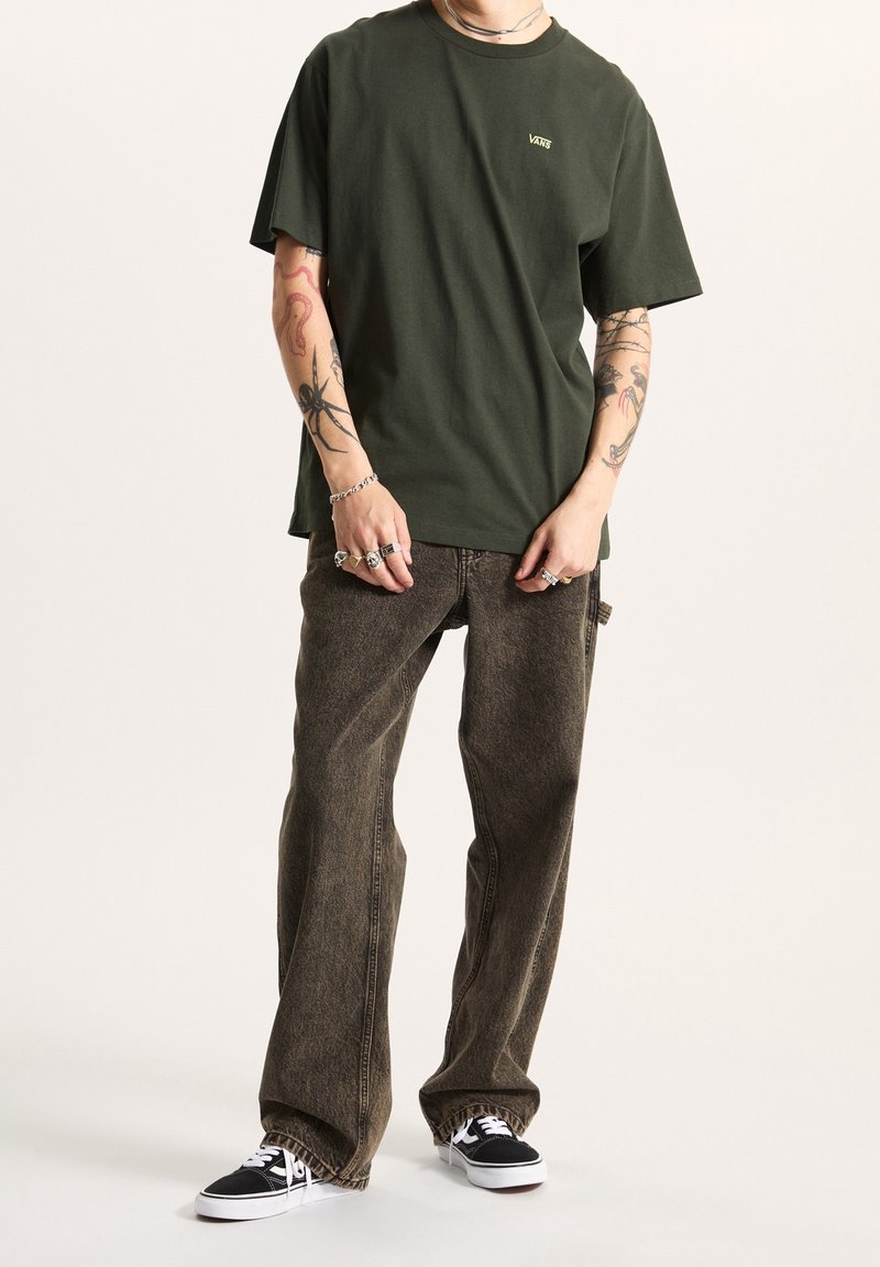 Personne portant un t-shirt Vans vert foncé, un jean marron ample, des baskets Vans noires, des tatouages visibles sur les bras, ainsi que des bagues et bracelets argentés.