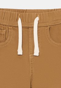 Pantalones marrones con cintura elástica y cordón blanco, costuras detalladas y bolsillos frontales visibles.