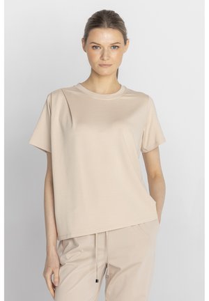 Kobieta z jasnobrązowymi włosami związanymi w kucyk, ubrana w beżowy t-shirt z krótkim rękawem i pasujące spodnie, stojąca na jednolitym tle.