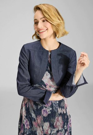 Kobieta z krótkimi blond włosami uśmiechająca się, ubrana w ciemnoniebieską krótką kurtkę na kwiecistej sukience z odcieniami fioletu i różu.