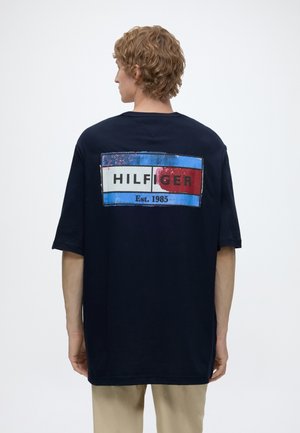 Młody mężczyzna z krótkimi kręconymi włosami, noszący granatowy, oversize'owy t-shirt z logo "Hilfiger Est. 1985" w kolorach czerwonym, białym i niebieskim na plecach.