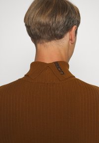 Maglione marrone a coste con collo alto e dettaglio con bottoni sul retro. Il tessuto ha una texture morbida con una forma aderente.