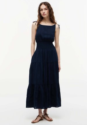 Jonge vrouw die een mouwloze navy maxi-jurk met laagjes en vastgeknoopte schouderbandjes draagt, samen met bruine platte sandalen, staand tegen een witte achtergrond.