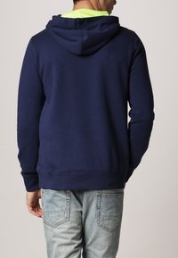 Dunkelblaues Kapuzensweatshirt mit geripptem Saum, aus Baumwollmaterial und mit einer weichen Textur. Sichtbare Nähte und kontrastierende neongrüne Innenauskleidung.