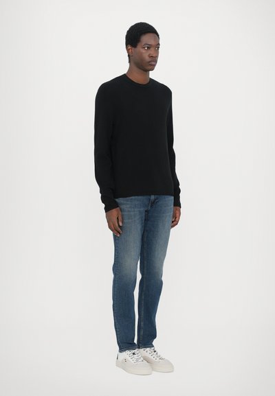 Pull en tricot noir à col rond, associé à un jean bleu clair et des baskets blanches. Le mannequin se tient de profil devant un fond uni.