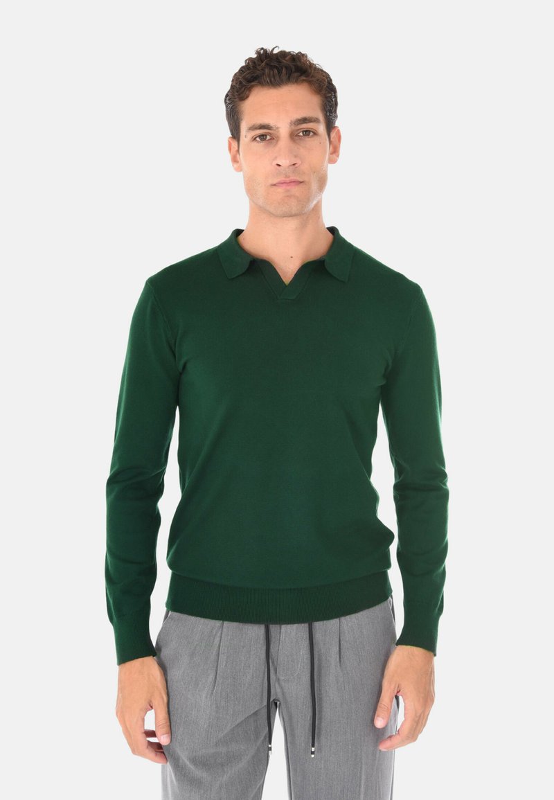 Pullover verde con maniche lunghe, scollatura a V e colletto. Tessuto morbido e design aderente. Indossato con pantaloni a pieghe grigi chiari.