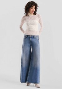 Weite blaue Jeans mit einem hellen Waschung, ausgestattet mit einem hohen Bund und Gesäßtaschen. Kombiniert mit einem transparenten weißen Spitzenoberteil mit langen Ärmeln.