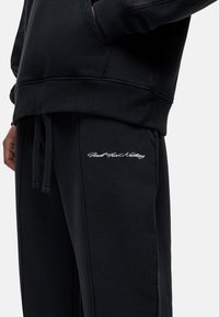 Zwarte sweatshirt en bijpassende joggers, gemaakt van zachte stof. Kenmerkt zich door een kangoeroezak en geborduurde tekstdetails op de jogger.