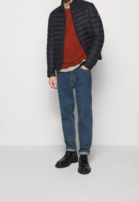 Puchowa kurtka w czarnym kolorze, teksturowana powierzchnia; sweter w kolorze rdzy; jasna koszula; niebieskie dżinsy; czarne buty na sznurówki z gumową podeszwą.