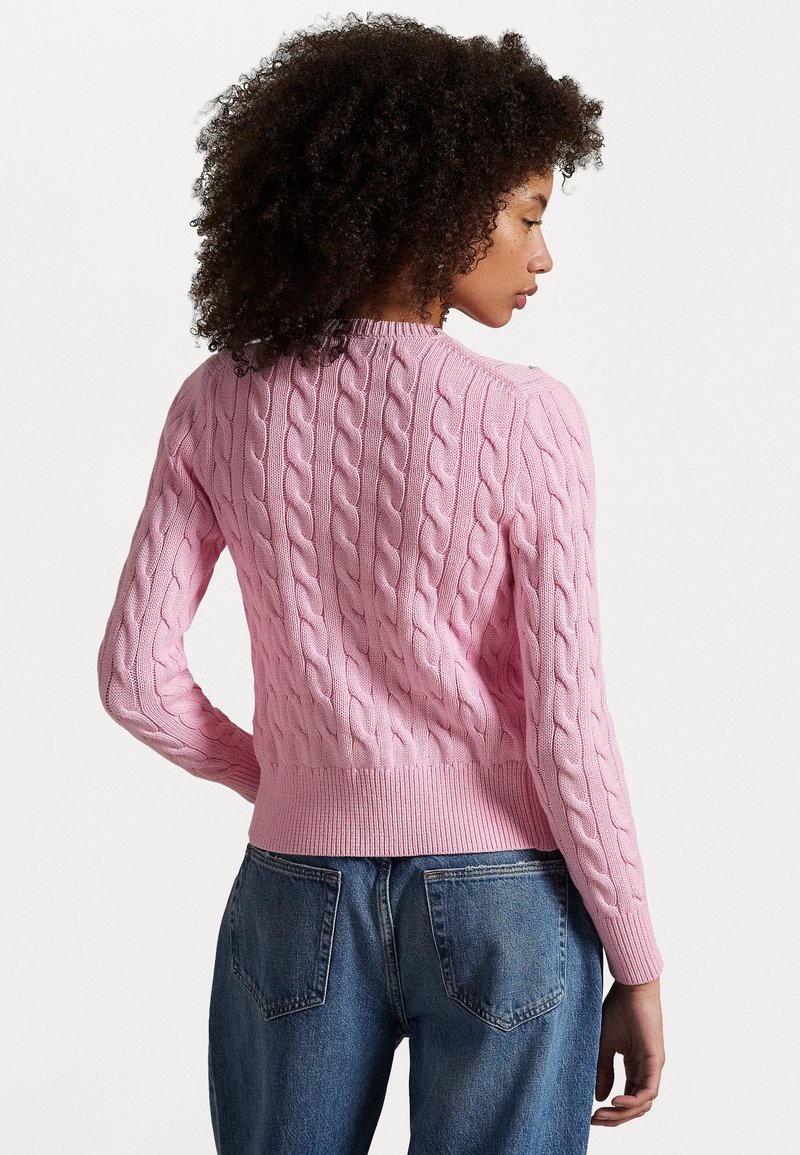 Woman with curly hair wearing a fitted pink cable-knit sweater and blue jeans, facing sideways against a plain background.