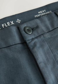 Zsúfolt kép sötétszürke nadrágról, gombos záródással és látható belső derékrész felirattal, ami a "FLEX" és a "NEXT PERFORMANCE" szavakat tartalmazza.