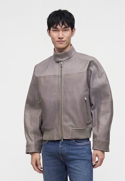 Jeune homme portant une veste en cuir grise avec un col montant et un jean bleu, debout devant un fond clair et uni.