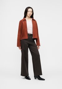 Vrouw draagt een roestkleurige jas met rits, lichtroze shirt, zwarte wijde broekspijpen, zwarte laarzen met hak, staand tegen een effen achtergrond.