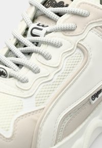 Baskets blanches et beige avec des lacets à motifs, des panneaux en mesh et une étiquette de marque sur le côté.