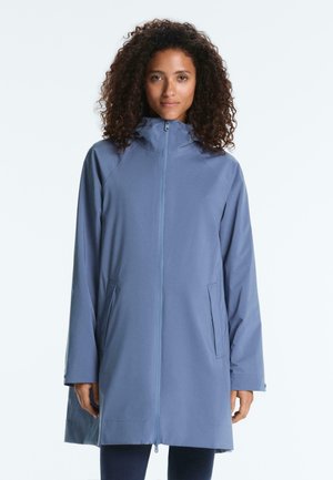 Kvinde med krøller iført en lyseblå hættetrøje med lynlås foran og sidelommer, stående foran en ensfarvet baggrund.