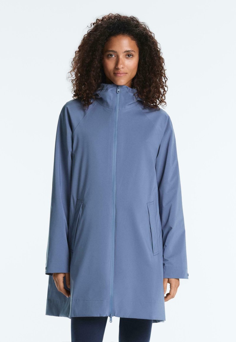 Femme aux cheveux bouclés portant un manteau à capuche bleu clair avec une fermeture éclair frontale et des poches latérales, debout devant un fond uni.