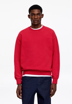 Młody mężczyzna stoi, ubrany w czerwoną długą bluzę na białą koszulę, w połączeniu z ciemnoniebieskimi spodniami, na jednolitym białym tle.