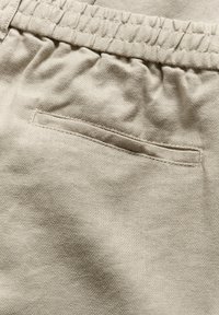 Beige hlače iz tkanine s elastičnim pasom in enim zadnjim žepom. Material ima teksturirano tkanje. Oblika je preprosta in funkcionalna.