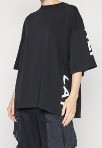 Černé oversize tričko, bavlněná tkanina, kulatý výstřih, krátké rukávy, bílé písmo na levém rukávu. Uvolněný střih, rovný lem.