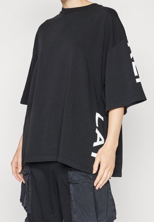 T-shirt nera oversize, in tessuto di cotone, collo rotondo, maniche corte, con testo bianco sulla manica sinistra. Vestibilità rilassata, orlo dritto.