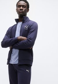 Μπλε ναυτικό αθλητικό μπουφάν με φερμουάρ και όρθιο γιακά, συνδυασμένο με ανοιχτό μπλε πουκάμισο και μπλε παντελόνι. Διαθέτει λευκή λεπτομέρεια λογότυπου στο μπουφάν.