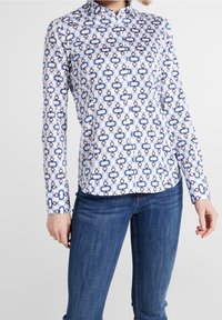 Bílá košile s tlačítky, zdobená modro-námořnickým vzorem připomínajícím řetěz. Má límec, dlouhé rukávy, přiléhavý střih a hladkou bavlněnou texturu.