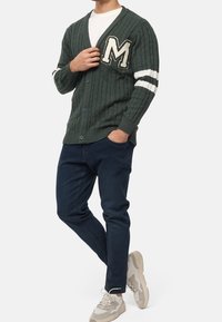 Cardigan verde a maglia intrecciata con scollo a V, caratterizzato da strisce crema e una toppa con la lettera 'M', abbinato a jeans blu scuro e sneakers beige.